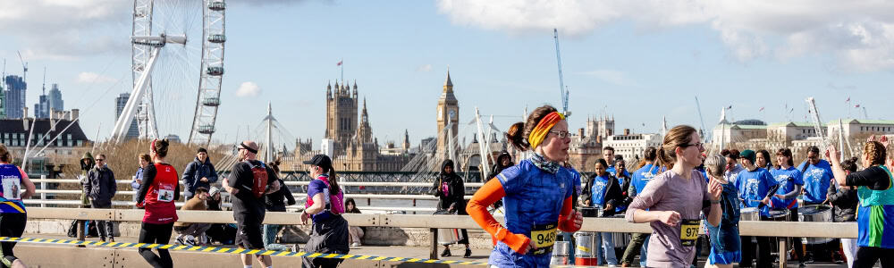 People running across a bridge in London with the London eye and parliment building in the background.